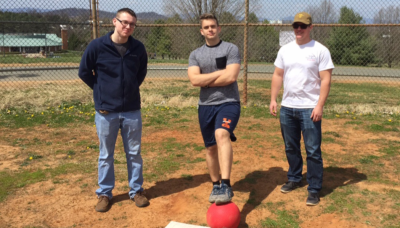 Three students stand on field. One has his foot on a kickball. 