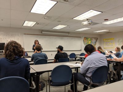 Students are taught Geology in a large classroom. The professor stands in the front of the class and students listen. 