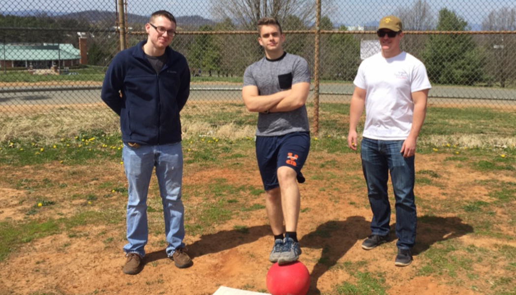 Three students stand on field. One has his foot on a kickball.