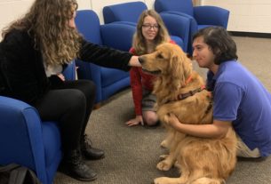 Two students sit with dog Maker's Joy on the floor. One pats the dog.
