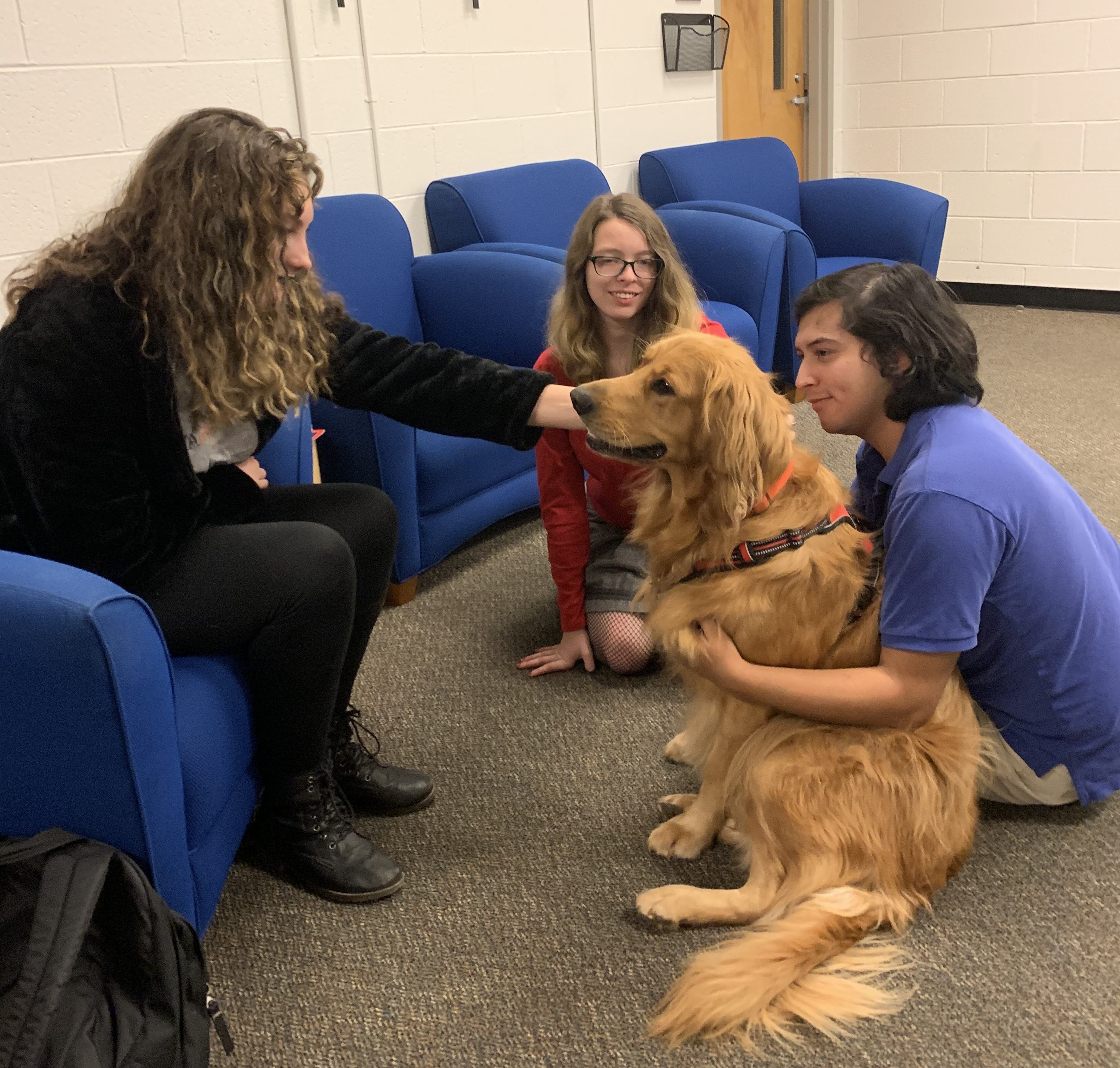 Two students sit with dog Maker's Joy on the floor. One pats the dog.