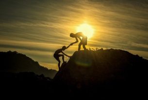 Two people climbing a mountain, one helping the other while the sun shines in the background