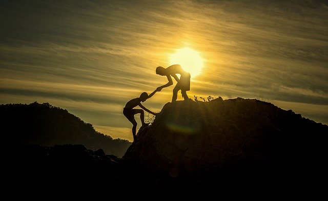 Two people climbing a mountain, one helping the other while the sun shines in the background