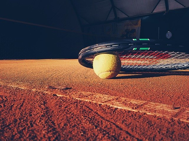 Angled picture of a tennis racket resting on a tennis ball on a clay court.