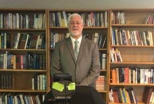 Tony Monaghan standing in front of wall lined with bookshelves. He is wearing a gray suit. The webcam he is using to stream his services can be seen.