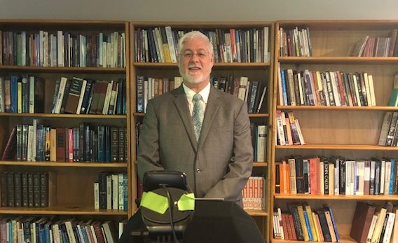 Tony Monaghan standing in front of wall lined with bookshelves. He is wearing a gray suit. The webcam he is using to stream his services can be seen.