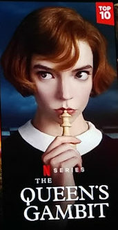 a photo of the queen's gambit netflix poster, a young redheaded woman (anna taylor-joy) holds a chees piece to her lips and looks thoughtfully to the left.