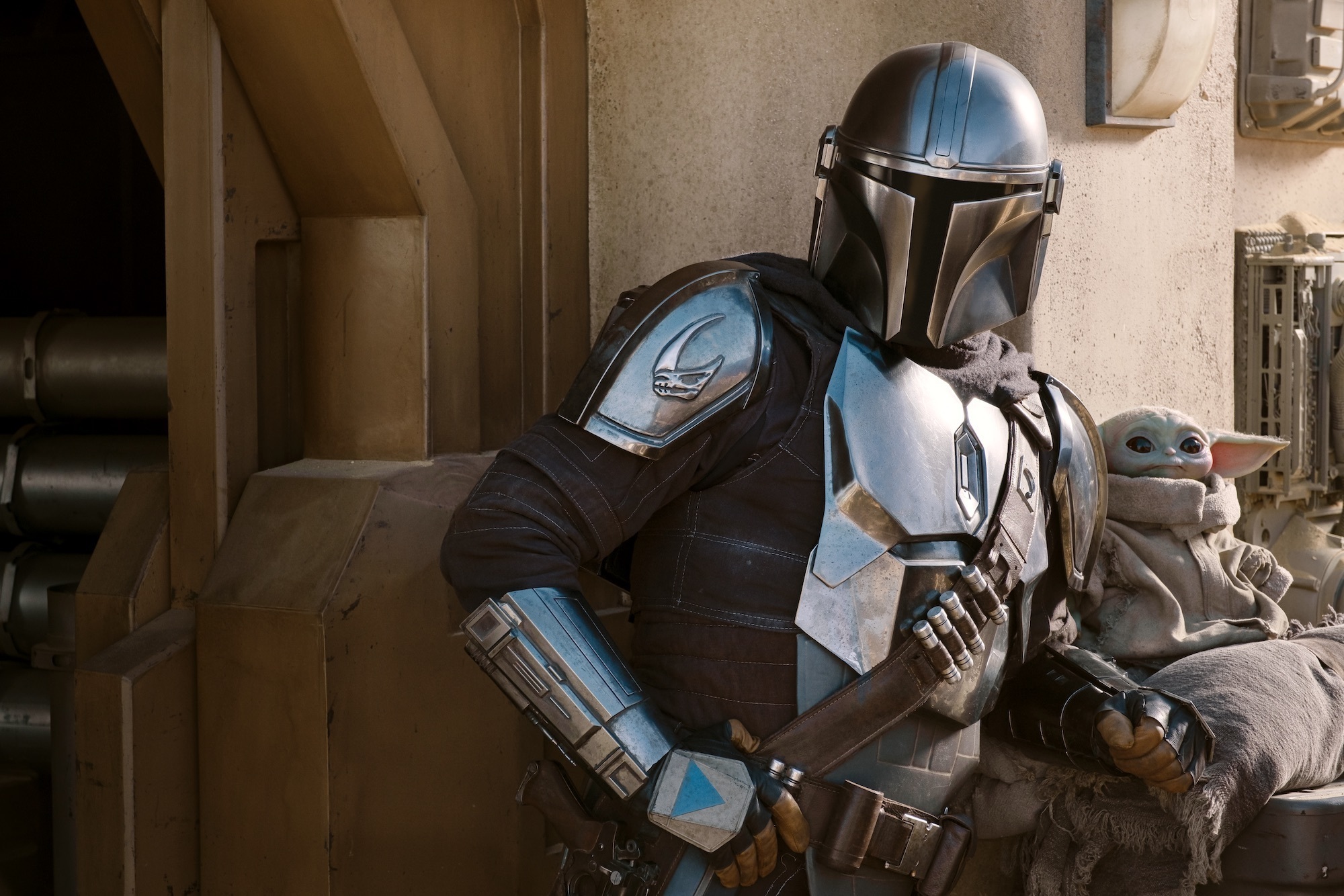 The Mandalorian leans against his speeder, where The Child is perched. They are in a desert town, The Mandalorian faces towards the camera while The Child looks offscreen to the right.