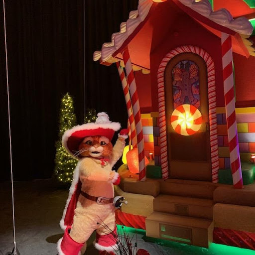 Puss in Boots in a festive outfit gestures towards a gingerbread house