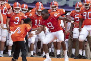 A football player clad in an orange jersey dances with a child while other football players stand in the background