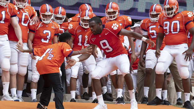 A football player clad in an orange jersey dances with a child while other football players stand in the background