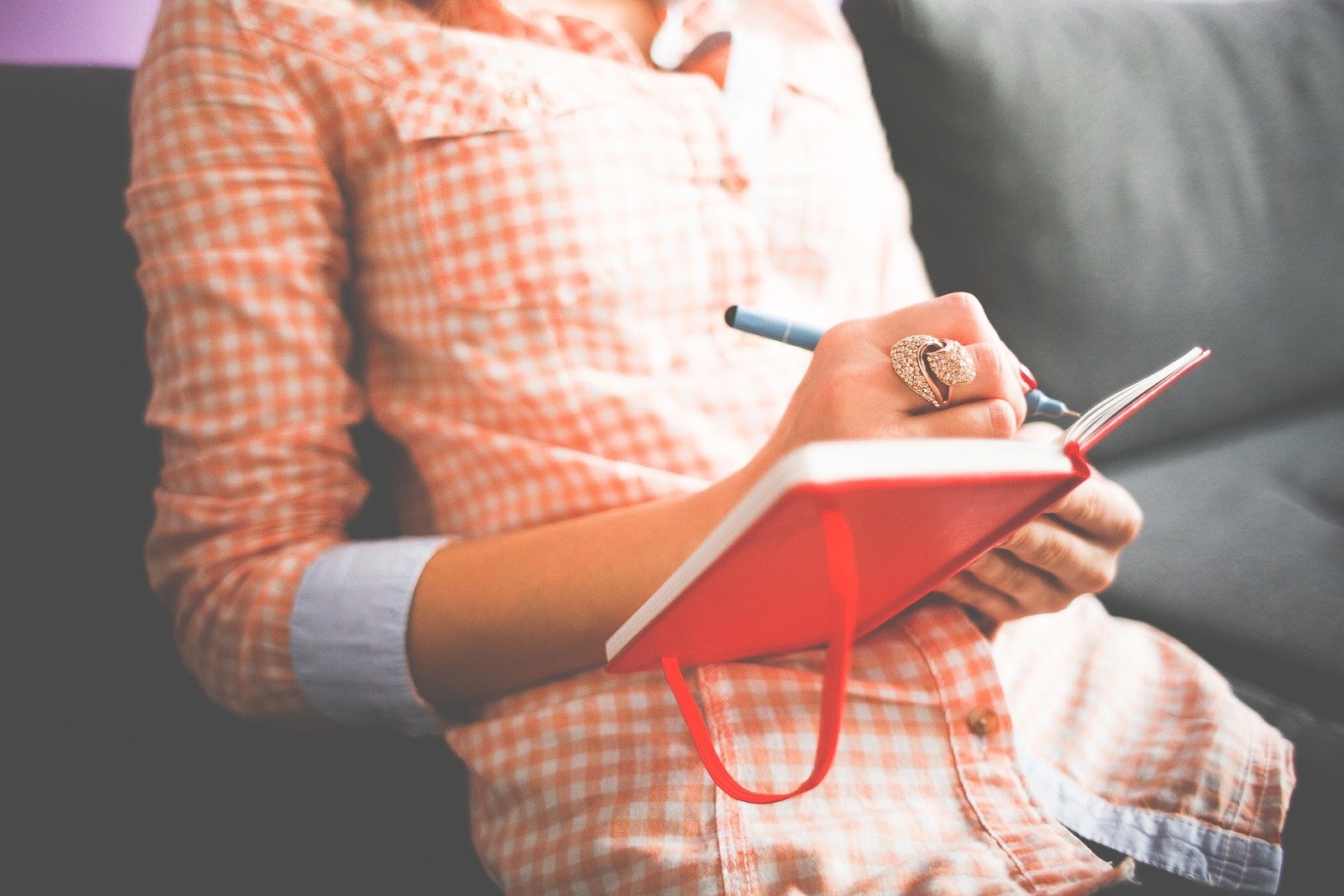 A woman sits on a sofa reclining and writing in a red-colored journal.