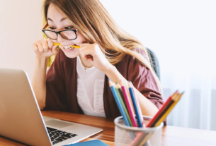 A woman looks at her computer and bites a pencil with a stressed look on her face.