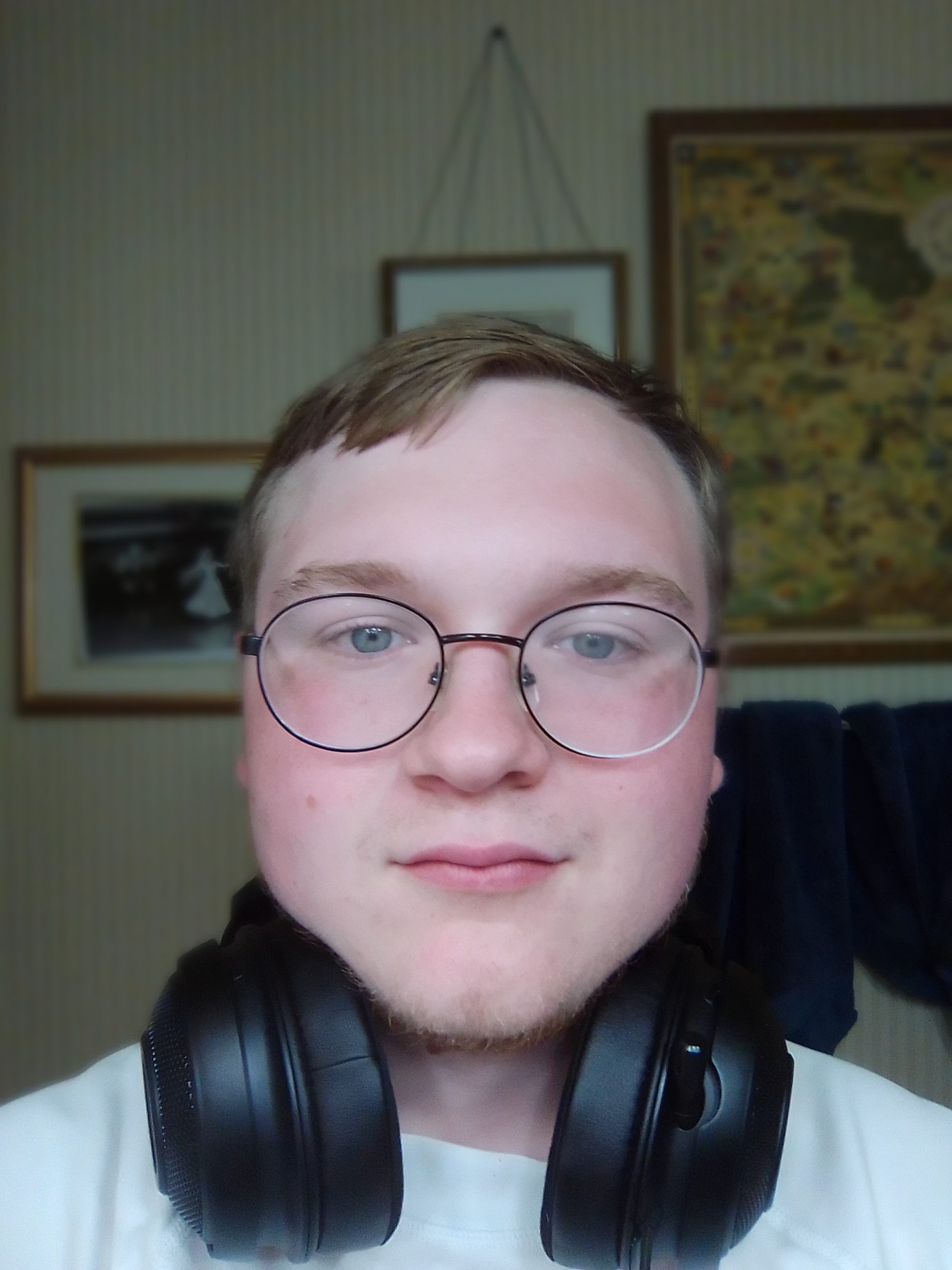 A man wearing headphones around his neck with brown hair and black rimmed glasses looks at the camera