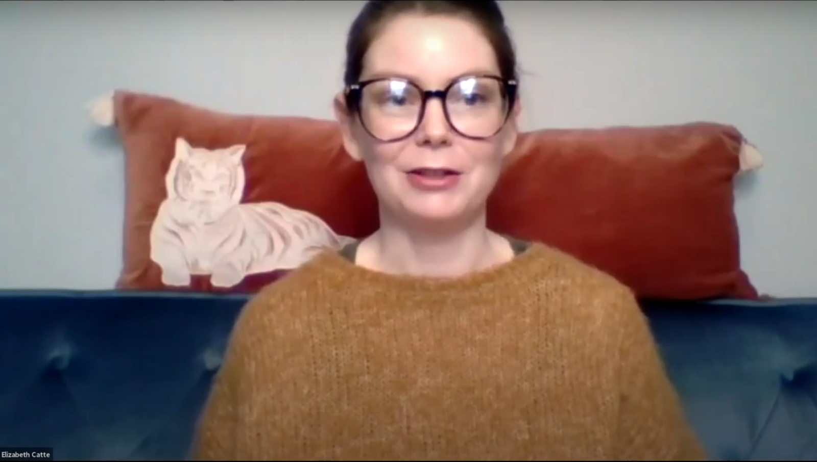 A woman with brown hair, a comfy-looking yellow sweater and black rimmed glasses sits facing the camera