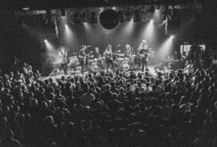 In a black and white photo, the members of King Gizzard perform live at a concert surrounded by a crowd of their fans