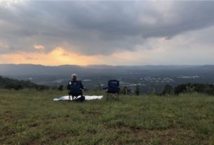 Man sits on the lawn of Carter Mountain, overlooking the city below and the sunset.