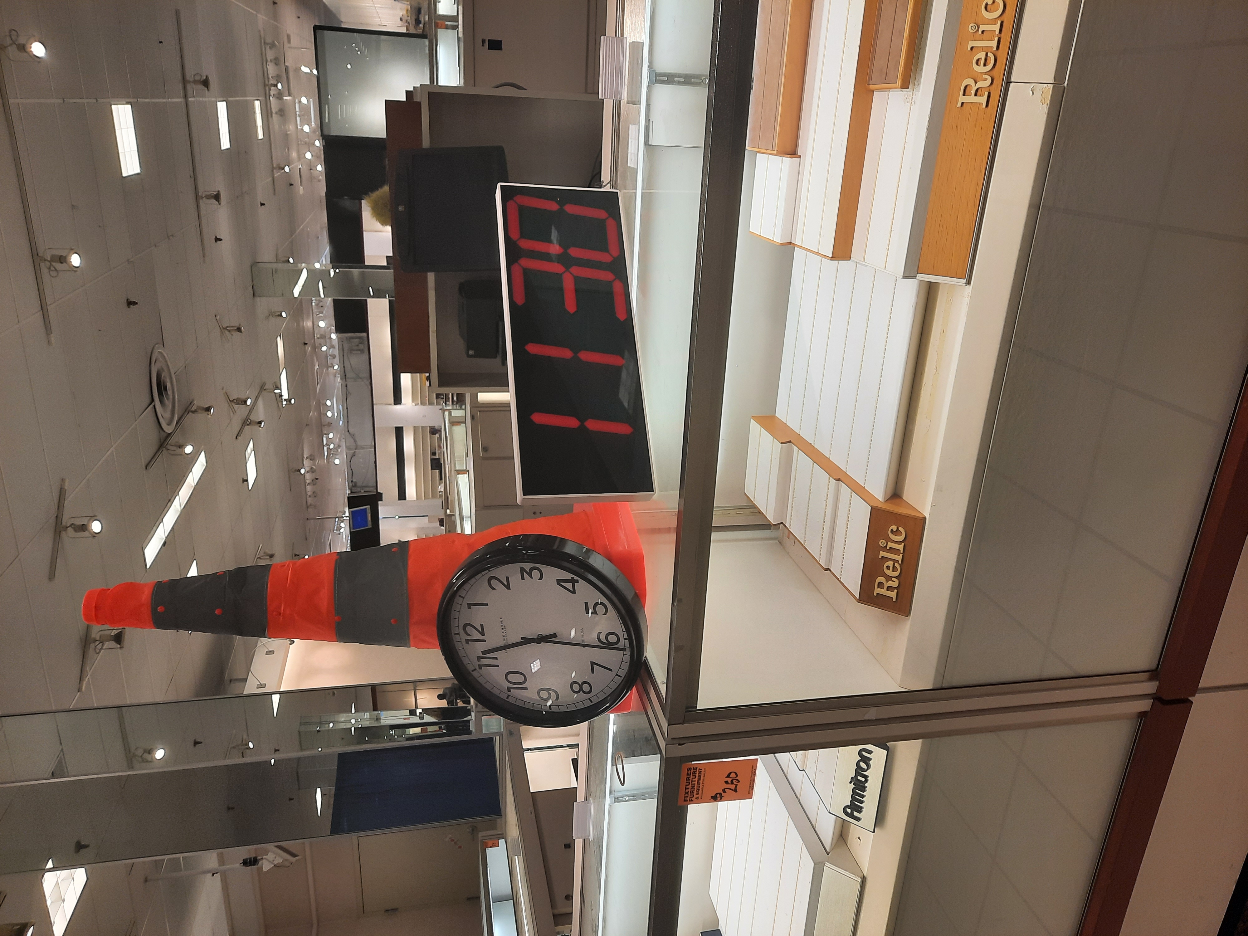 An analogue clock sits propped up against a bright orange traffic cone, and a digital clock sits next to it. Both clocks display the time 11:30