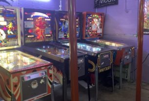 A row of colorful pinball machines sit side by side in a room with a concrete floor and purple walls