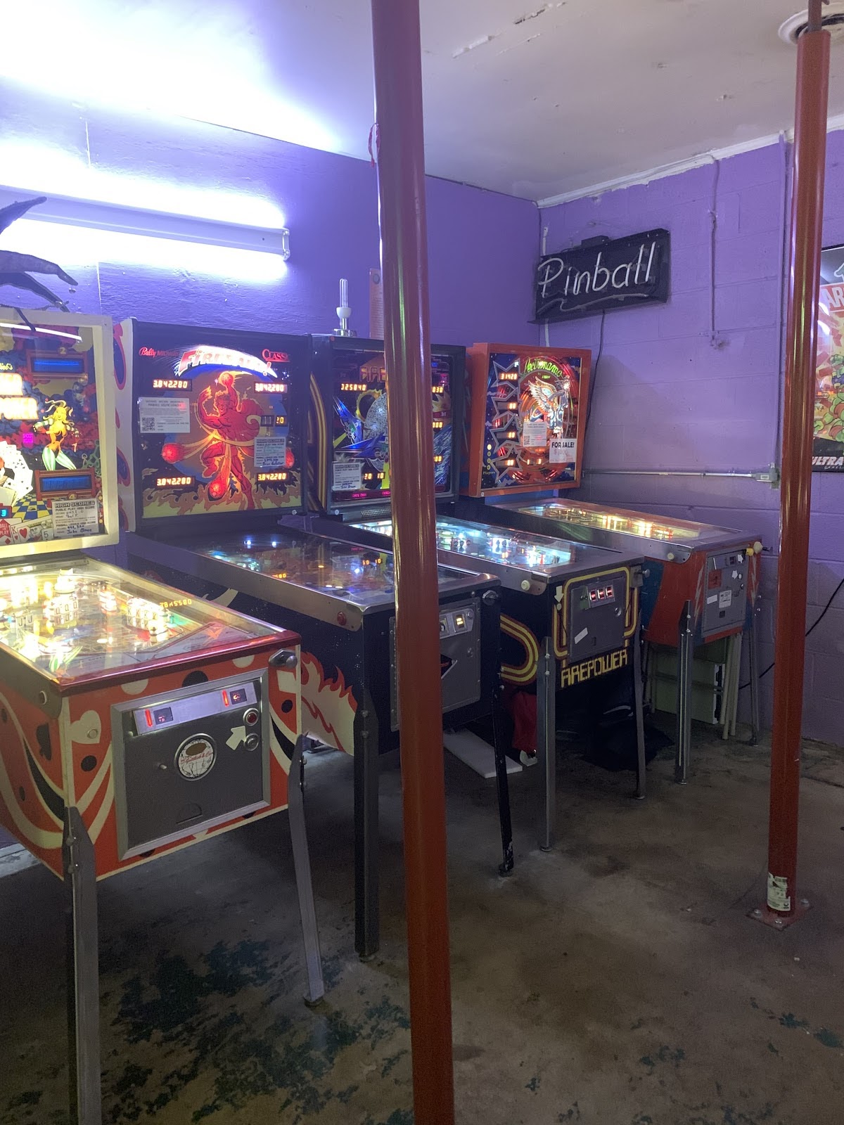 A row of colorful pinball machines sit side by side in a room with a concrete floor and purple walls