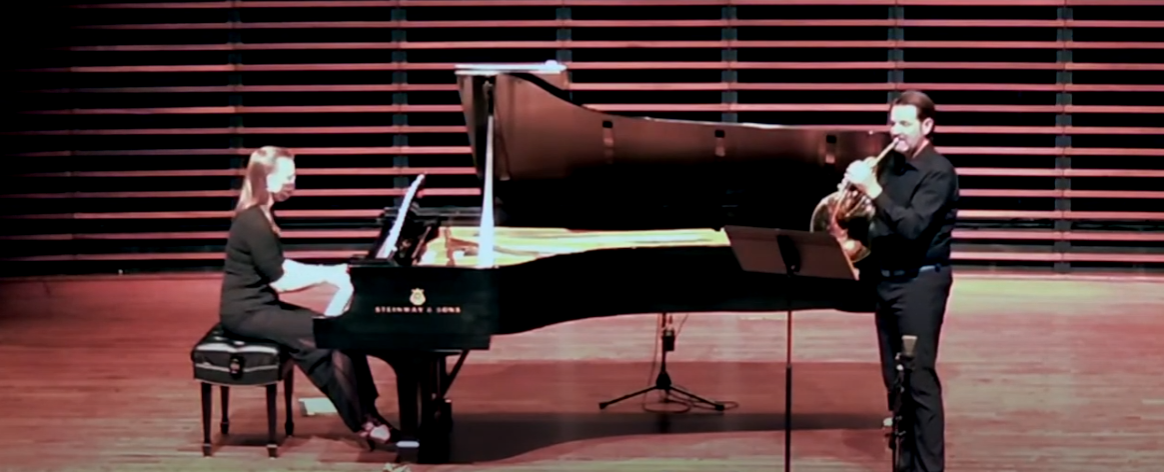 Tracey Schimmel-Reed plays piano on a stage while Ian Zook plays the french horn