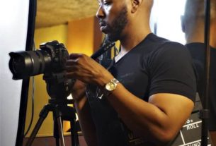 Ty Cooper stands facing left looking in his camera while holding a film clapboard