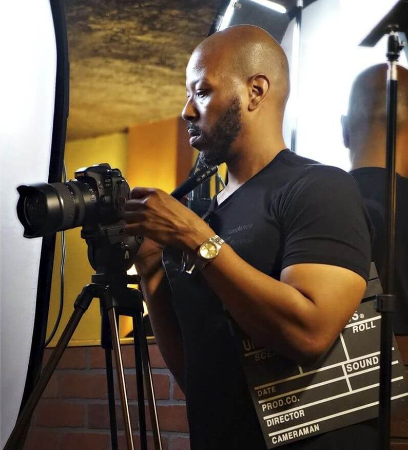 Ty Cooper stands facing left looking in his camera while holding a film clapboard