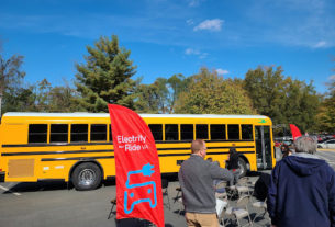 Several people stand in a parking lot next to a yellow electric school bus, next to a red banner reading "Electrify your ride VA" in white text with a blue picture of an extension cord