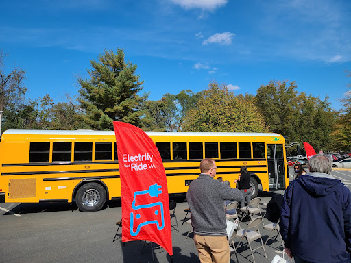 Several people stand in a parking lot next to a yellow electric school bus, next to a red banner reading "Electrify your ride VA" in white text with a blue picture of an extension cord