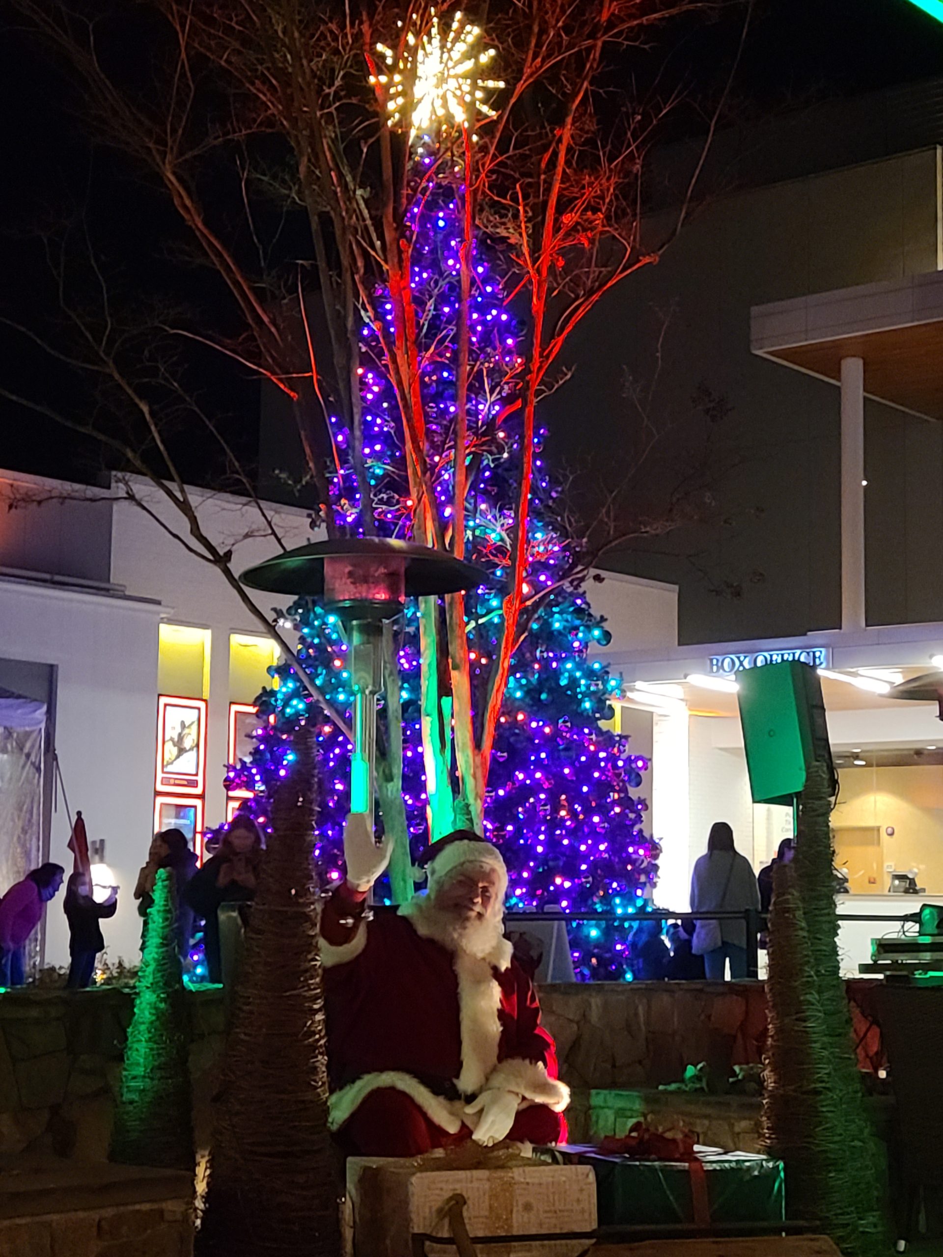 Santa Claus poses in front of a large Christmas Tree lit with red, blue and green lights, with a white star on top