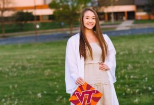 A young woman in a white dress stands on a green lawn holding an orange and red graduation cap with the letters "VT" on it in red