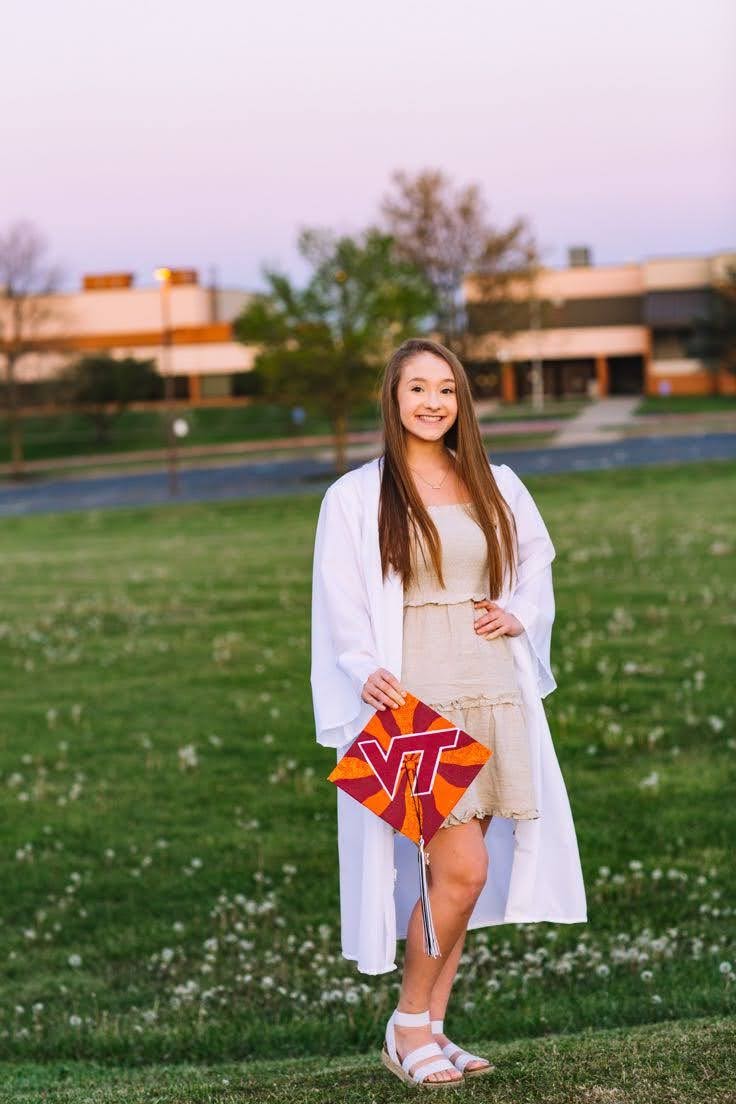 A young woman in a white dress stands on a green lawn holding an orange and red graduation cap with the letters "VT" on it in red