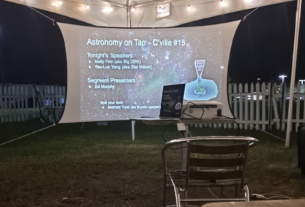 Outside under a tent with string lights strung across the top, is a projector screen projecting a slideshow with the background being an image of a picture taken of the stars in space. The projector screen states "Astronomy on Tap," with a list of the guest speakers.