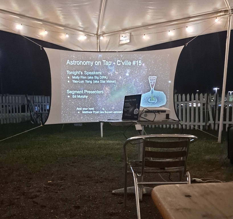 Outside under a tent with string lights strung across the top, is a projector screen projecting a slideshow with the background being an image of a picture taken of the stars in space. The projector screen states "Astronomy on Tap," with a list of the guest speakers.