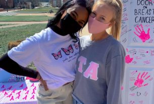 two women pose for a photo on a sunny day, behind them a white posterboard with pink handprints on it reads "slamming the door on breast cancer"