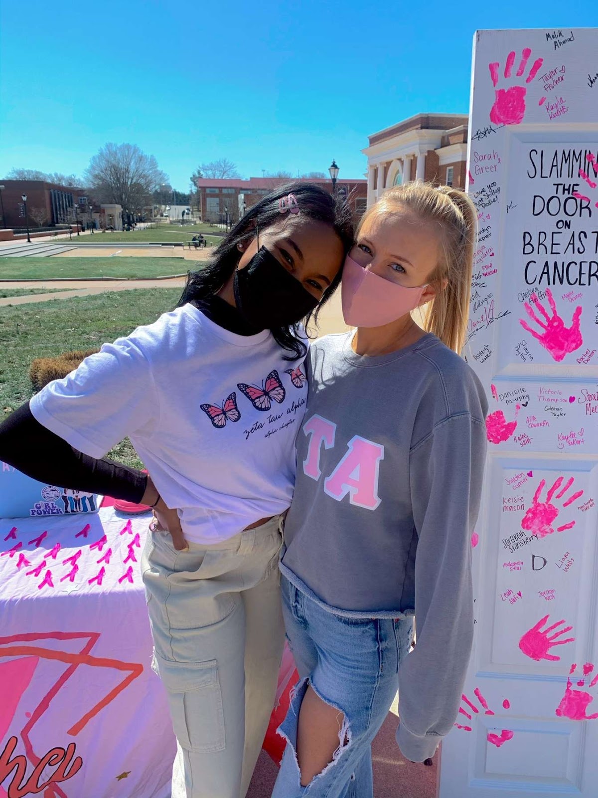 two women pose for a photo on a sunny day, behind them a white posterboard with pink handprints on it reads "slamming the door on breast cancer"