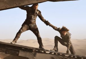 An older bearded man holds on to a younger man as they both balance on the ramp of an aircraft with a sand dune in the background