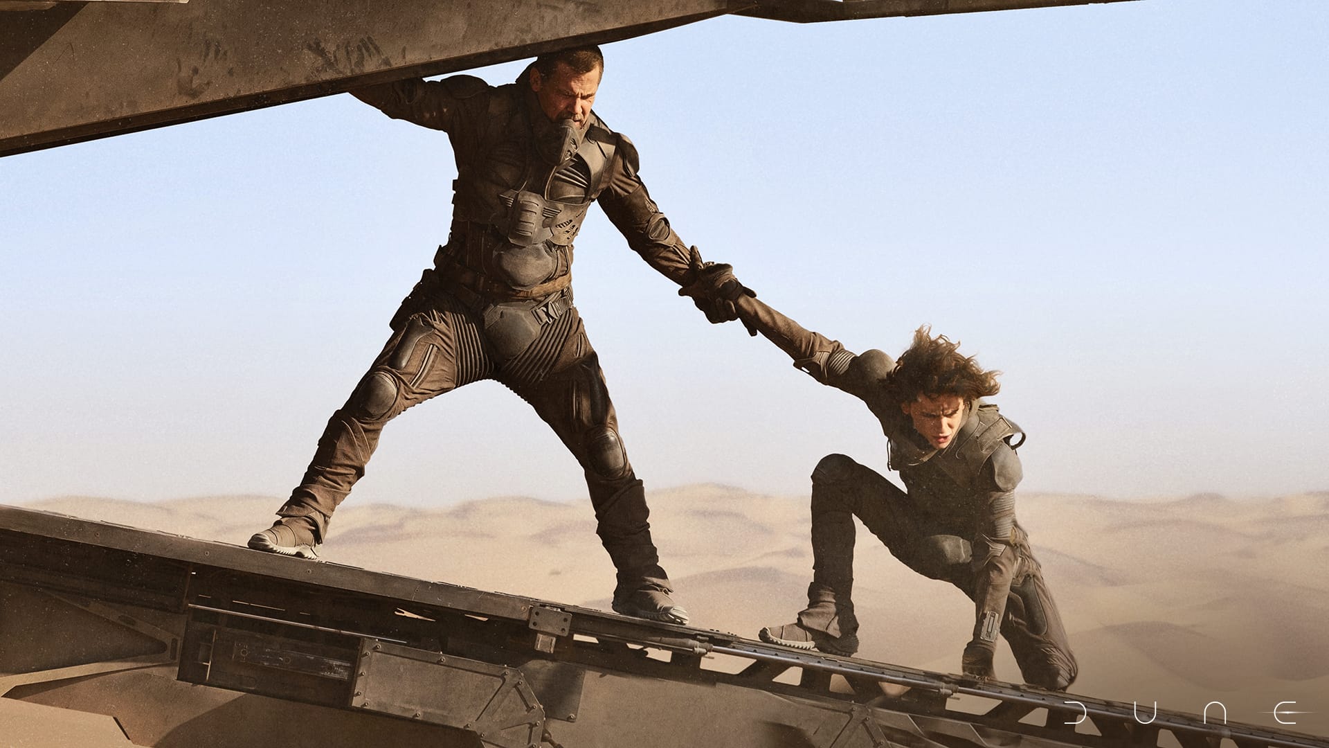 An older bearded man holds on to a younger man as they both balance on the ramp of an aircraft with a sand dune in the background