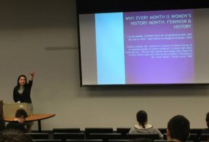 Woman standing in a lecture hall, with arm outstretched making a dropping an item expression with her hand. Projector screen in the background with text that reads "Why every month is women's history month."