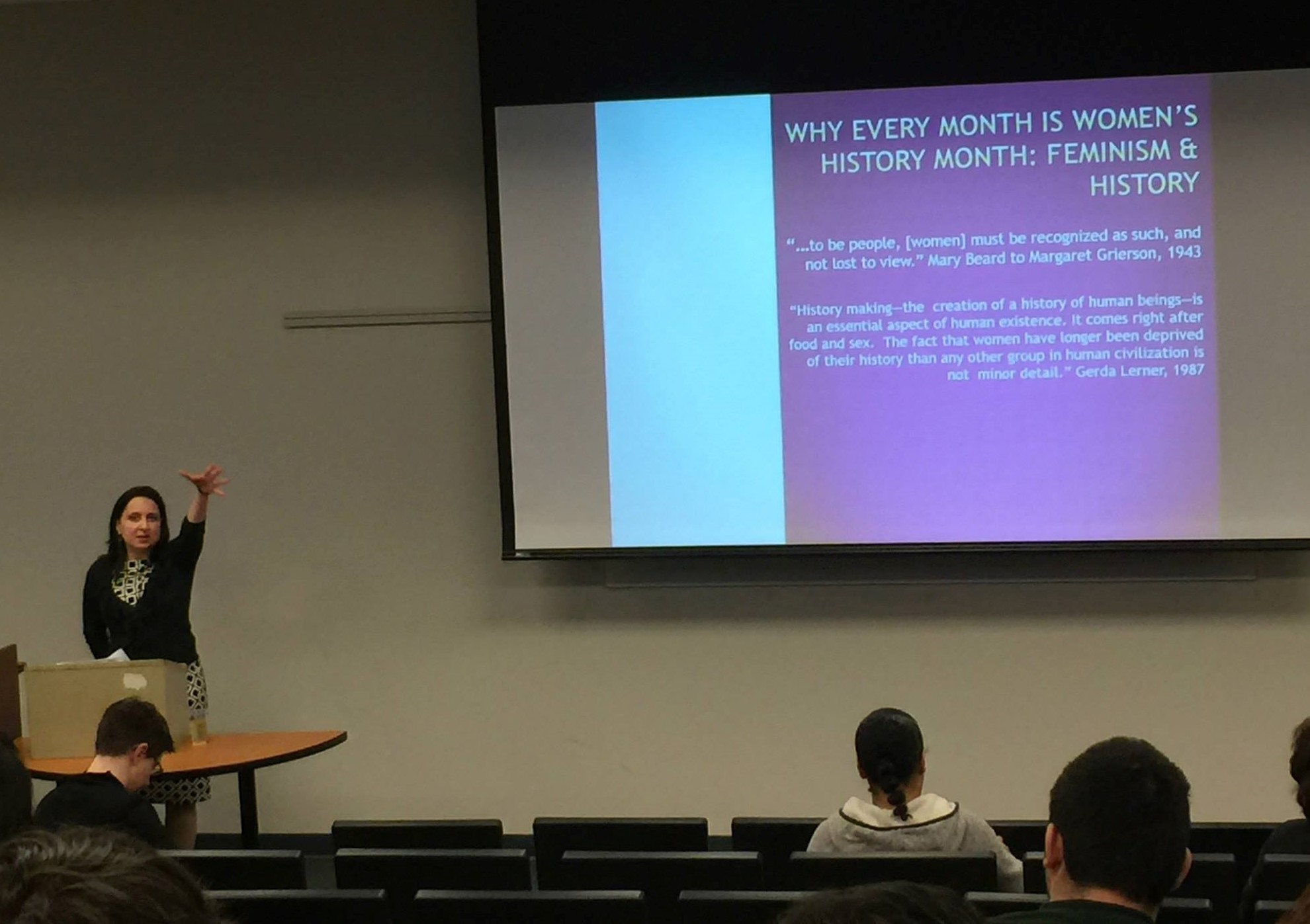 Woman standing in a lecture hall, with arm outstretched making a dropping an item expression with her hand. Projector screen in the background with text that reads "Why every month is women's history month."