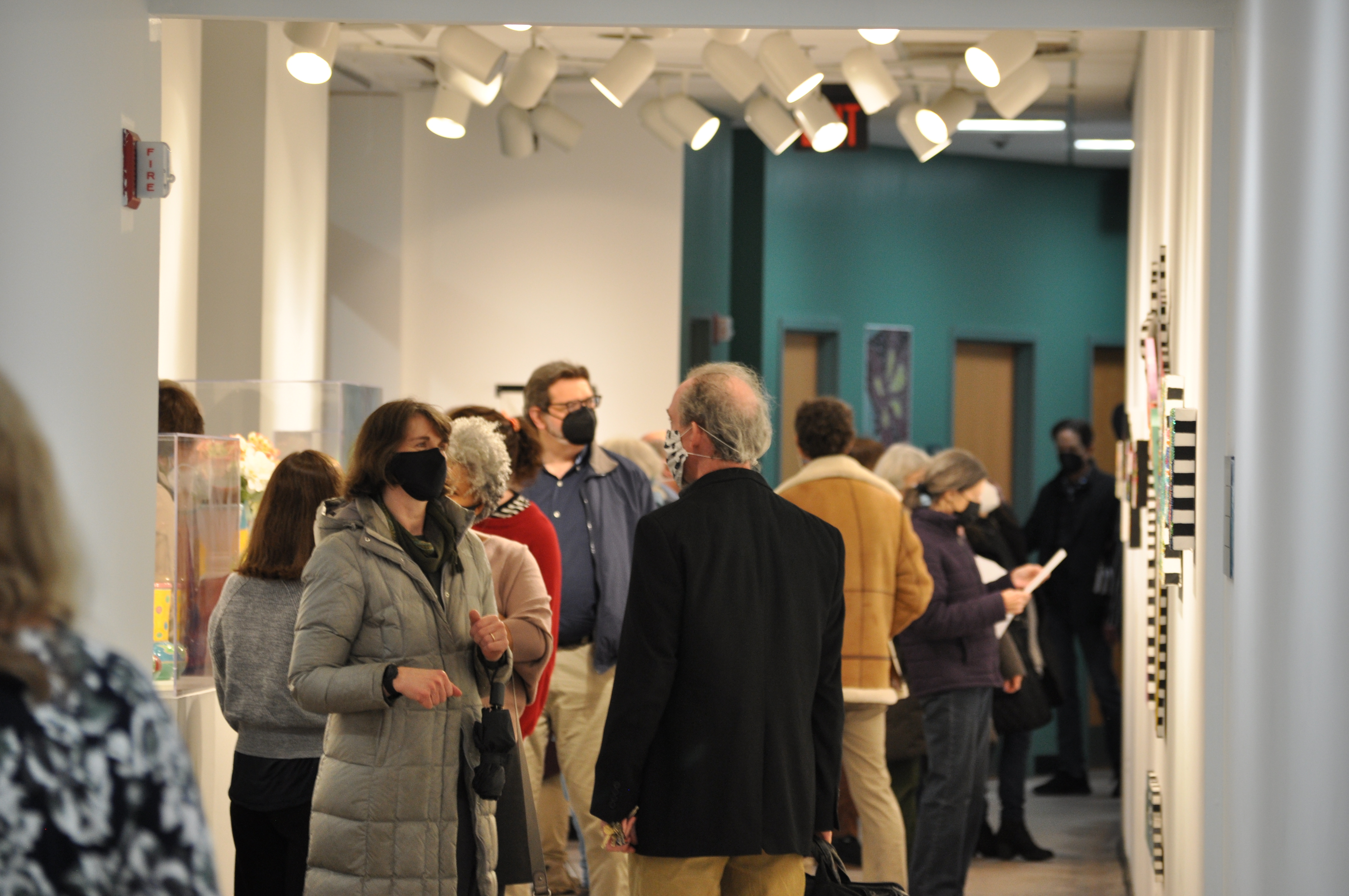 Crowded hallway with an array of lights beaming on the walls, which are adorned with paintings and art. The back wall of the hallway is teal, with a door with paintings on either side of it.