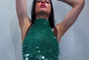 Arca posing in a shiny greenish blue outfit.