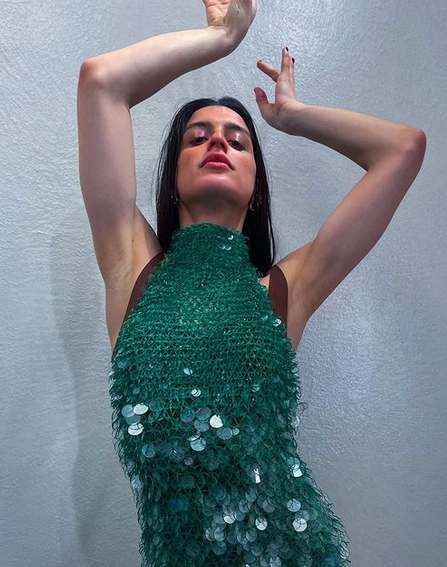 Arca posing in a shiny greenish blue outfit.