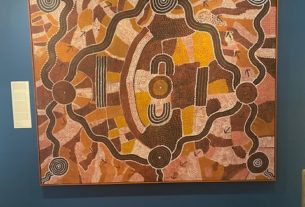 aboriginal art on display on a blue wall, over a stained hardwood floor. The palette of the painting is heavy on brown and yellow colors, and the art features wavy lines and blocks made up of dots.