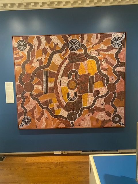 aboriginal art on display on a blue wall, over a stained hardwood floor. The palette of the painting is heavy on brown and yellow colors, and the art features wavy lines and blocks made up of dots.