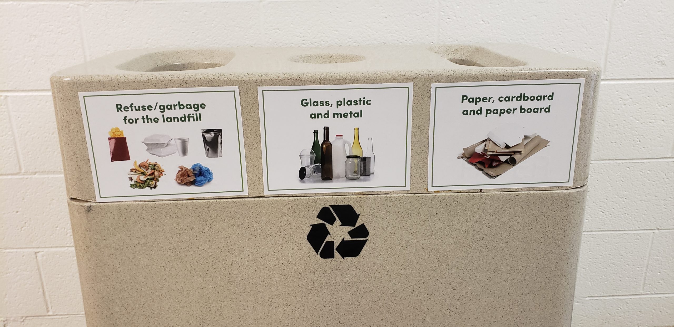 Recycling bin against a white cinderblock, with pictures of what can be recycled on each section of the bins.