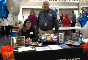 A woman sits and a man stands behind the RMC Events table. A banner for the PVCC Annual Job Fair hangs in the background.