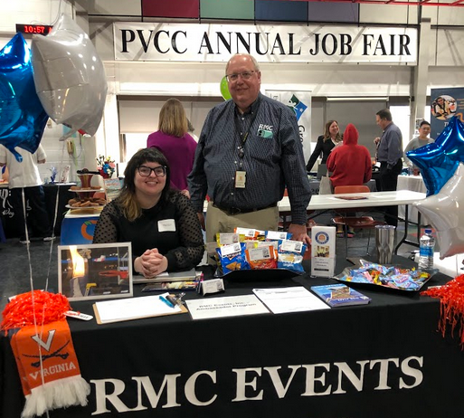 A woman sits and a man stands behind the RMC Events table. A banner for the PVCC Annual Job Fair hangs in the background.