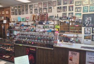 Tobacconist and Gifts main counter/register. On the back wall are pictures of various celebrities from the mid to late 20th century. On back wall are various pipes and tobacco supplies, such as pipe cleaners, cigar cutters, and novelty lighters. Off screen to the right is a register. On the far left of the counter is a glass display case with various pipes. To its right are 3 rows of jars filled with different flavors of pipe tobacco, with a scale to their immediate right for weighing the number of ounces of pipe tobacco are being purchased.