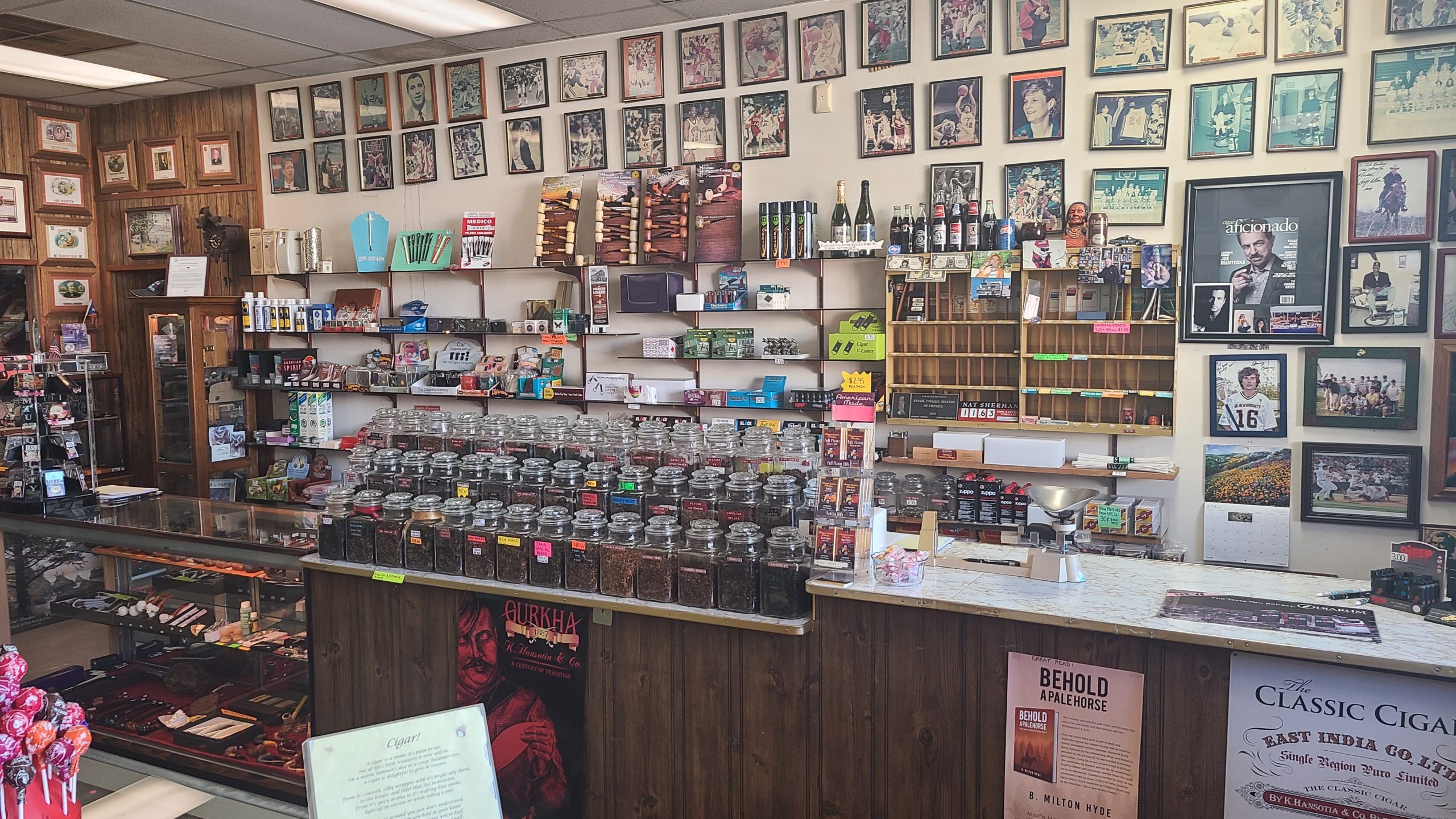 Tobacconist and Gifts main counter/register. On the back wall are pictures of various celebrities from the mid to late 20th century. On back wall are various pipes and tobacco supplies, such as pipe cleaners, cigar cutters, and novelty lighters. Off screen to the right is a register. On the far left of the counter is a glass display case with various pipes. To its right are 3 rows of jars filled with different flavors of pipe tobacco, with a scale to their immediate right for weighing the number of ounces of pipe tobacco are being purchased.